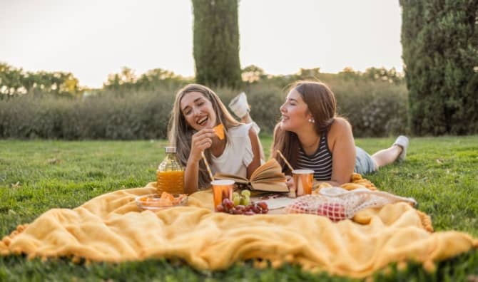 Two young women lounging on a yellow blanket in a sunny park.