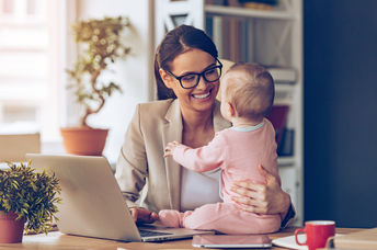 Cheerful young beautiful businesswoman looking at her baby girl with smile while sitting at her working place