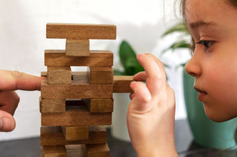 As a father and daughter play tumble tower, dad models resilience by assisting her in strengthening the tower.