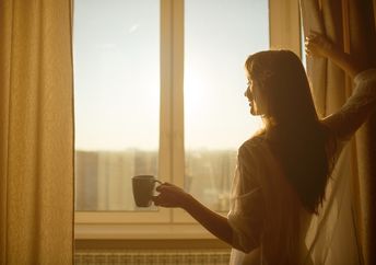 Woman in the morning. Attractive sexy woman with neat body is holding a cup with hot tea or coffee and looking at the sunrise standing near the window in her home and having a perfect cozy morning.