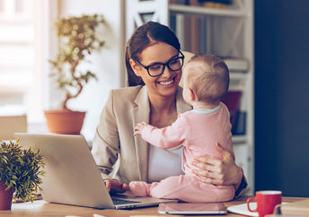 Cheerful young beautiful businesswoman looking at her baby girl with smile while sitting at her working place