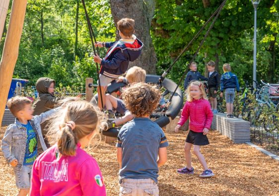 Elementary school children play outside in a playground with plenty of greenery.