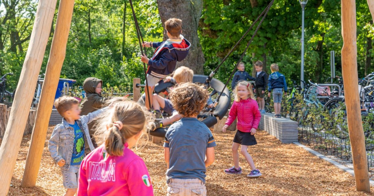 Elementary school children play outside in a playground with plenty of greenery.