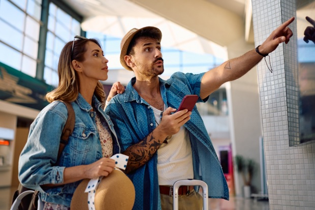A couple at an airport looks at a flight information screen while the man points toward it. 