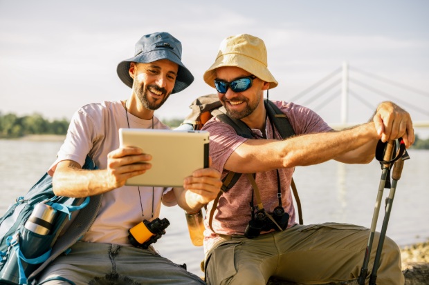 Two men resting on a fallen tree and viewing a tablet during a hiking trip. 