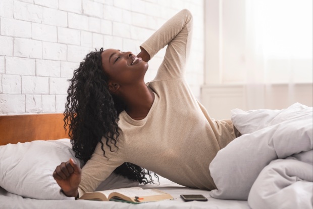 A woman stretches in bed with a book and phone next to her.