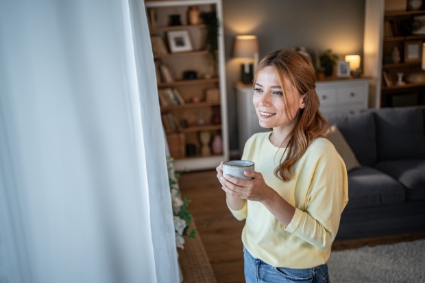 A woman stands by a window while holding a coffee cup.