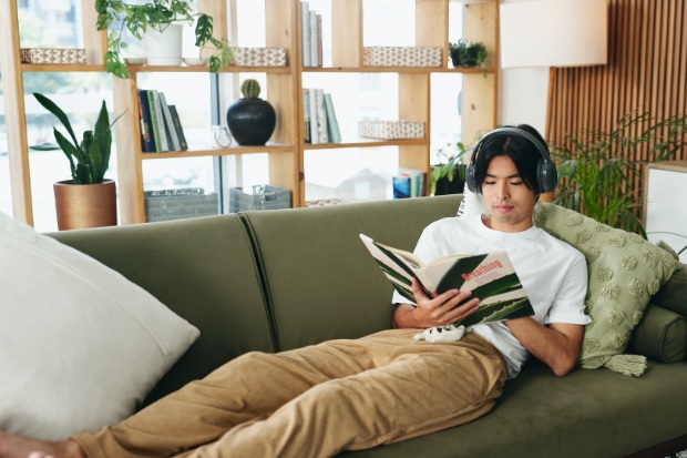 A man lays on a couch and reads while listening to music.